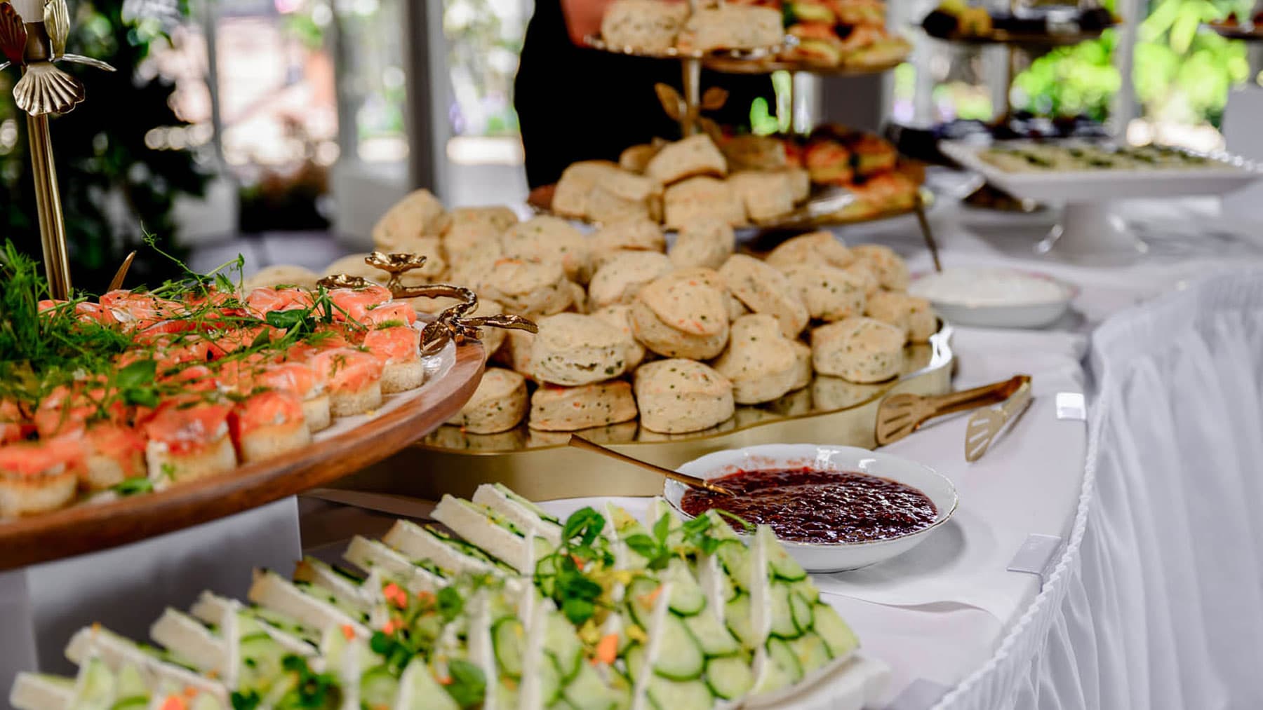 Buffet table with smoked salmon canapés, a pile of scones, jam, and cucumber sandwiches.